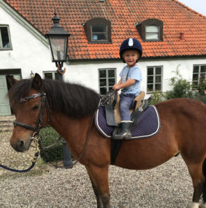 Ett litet barn sitter på en häst utanför ett av husen vid Grevlundagården